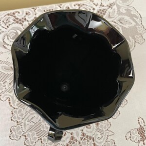 Peut inclure: Une vue plongeante sur un pichet en verre noir brillant dot&eacute; d'un large bord festonn&eacute; et ondul&eacute;. L'int&eacute;rieur est profond et sombre, r&eacute;v&eacute;lant une surface lisse et r&eacute;fl&eacute;chissante. Une petite partie d'une anse noire est visible en bas.