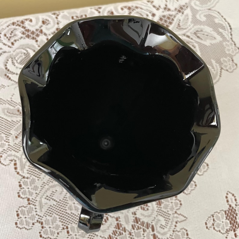 Peut inclure: Une vue plongeante sur un pichet en verre noir brillant dot&eacute; d'un large bord festonn&eacute; et ondul&eacute;. L'int&eacute;rieur est profond et sombre, r&eacute;v&eacute;lant une surface lisse et r&eacute;fl&eacute;chissante. Une petite partie d'une anse noire est visible en bas.