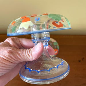 May include: A clear glass decorative piece with a mushroom-shaped top, adorned with colorful floral designs in orange, green, and blue. The base and rim feature blue painted accents. The piece is held by a hand.