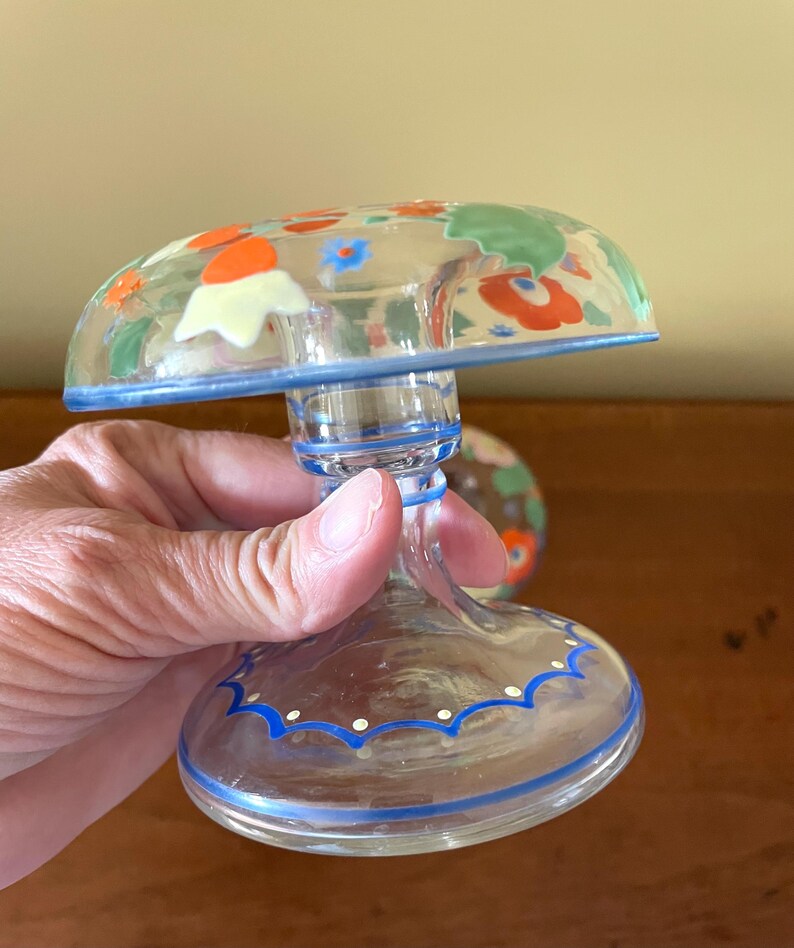 May include: A clear glass decorative piece with a mushroom-shaped top, adorned with colorful floral designs in orange, green, and blue. The base and rim feature blue painted accents. The piece is held by a hand.