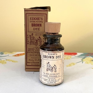May include: A glass bottle of Eddie's Everlasting Brown Dye with a cork stopper and a brown wooden cap. The bottle is labelled with the text "Eddie's Brown Dye" and instructions for use. The bottle is sitting on a white surface with a floral pattern.