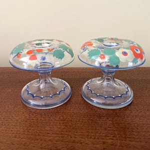 May include: Two clear glass candle holders with a floral design. The candle holders have a round top with a blue rim and a stem. The floral design includes red, white, and green flowers. The base of the candle holders has a blue scalloped design.