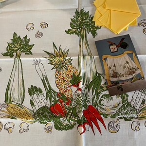 May include: A white tablecloth featuring a vibrant vegetable pattern with corn, pineapple, and red peppers. Yellow napkins are stacked on the right. A card with an apron design is also present.