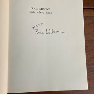May include: Open book page with the title "ERICA WILSON'S Embroidery Book" and an author's signature. The text is in a dark font on a white page. The book is open on a wooden surface.