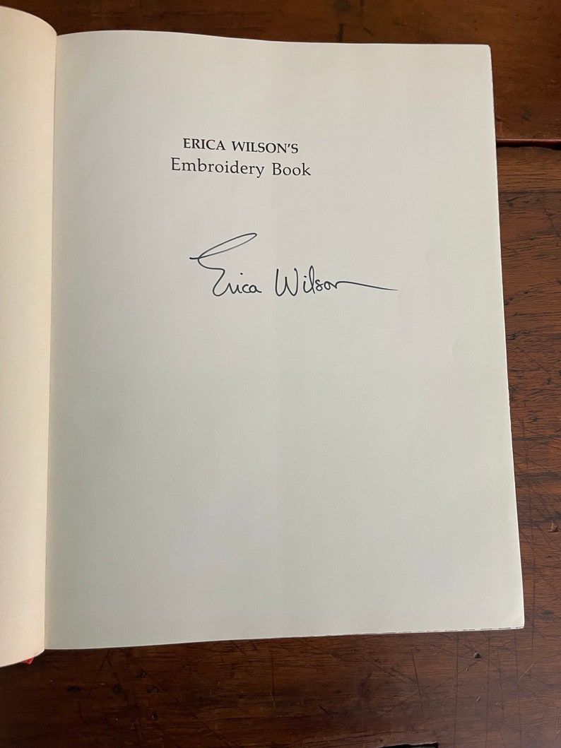 May include: Open book page with the title "ERICA WILSON'S Embroidery Book" and an author's signature. The text is in a dark font on a white page. The book is open on a wooden surface.