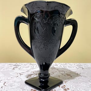 Peut inclure: Un vase en verre noir avec un bord &eacute;vas&eacute; et ondul&eacute; ainsi que deux anses lat&eacute;rales. Le corps pr&eacute;sente des figures en relief de deux personnes dansant dans un cadre en forme de c&oelig;ur, agr&eacute;ment&eacute; de volutes et d'une base carr&eacute;e.