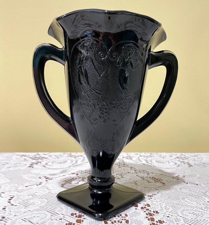 Peut inclure: Un vase en verre noir avec un bord &eacute;vas&eacute; et ondul&eacute; ainsi que deux anses lat&eacute;rales. Le corps pr&eacute;sente des figures en relief de deux personnes dansant dans un cadre en forme de c&oelig;ur, agr&eacute;ment&eacute; de volutes et d'une base carr&eacute;e.