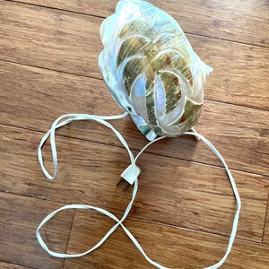 May include: A decorative seashell lamp with a carved palm tree design. The lamp has a pearlescent finish with brown and green accents. A white electrical cord and plug are attached.