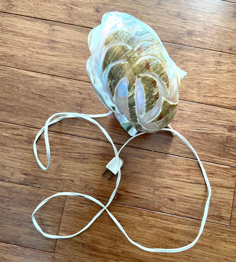 May include: A decorative seashell lamp with a carved palm tree design. The lamp has a pearlescent finish with brown and green accents. A white electrical cord and plug are attached.