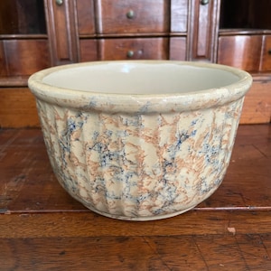 May include: A beige stoneware bowl with a ribbed exterior and a speckled pattern of blue and brown. The bowl has a wide, rounded shape and a smooth, off-white interior. A vintage kitchenware item.