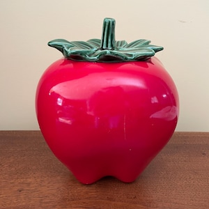 May include: A glossy red ceramic apple-shaped jar with a green leaf-shaped lid. The jar has a smooth, reflective surface and is designed to resemble a large apple. The lid features a stem.
