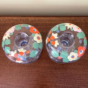 May include: Two clear glass candle holders with floral designs. The candle holders have a circular shape with a central opening for a candle. The floral design includes orange, red, yellow, and white flowers with green leaves.