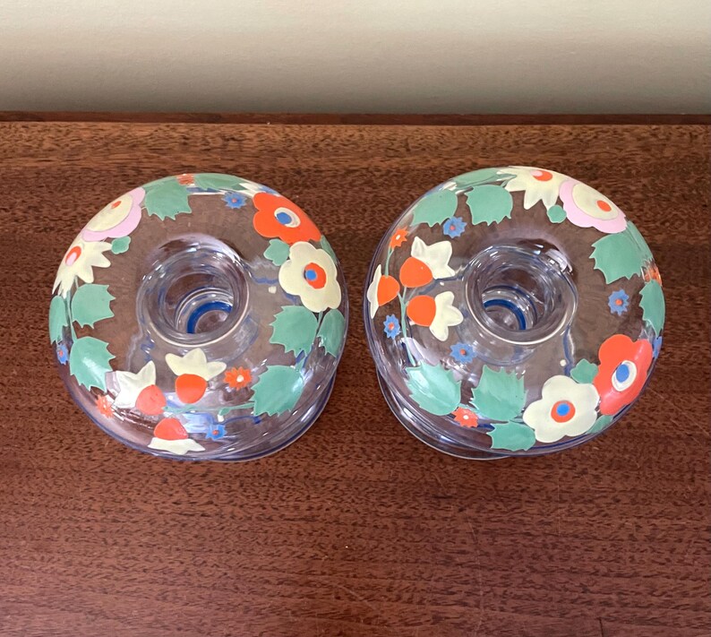 May include: Two clear glass candle holders with floral designs. The candle holders have a circular shape with a central opening for a candle. The floral design includes orange, red, yellow, and white flowers with green leaves.