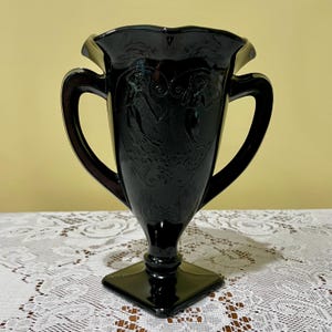 Peut inclure: Un vase en verre noir dot&eacute; de deux anses lat&eacute;rales, d'un bord festonn&eacute; et d'une base carr&eacute;e. Le corps du vase est orn&eacute; d'un motif en relief repr&eacute;sentant deux personnages et des volutes complexes sur la surface avant du verre sombre.