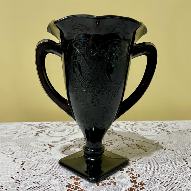 Peut inclure: Un vase en verre noir dot&eacute; de deux anses lat&eacute;rales, d'un bord festonn&eacute; et d'une base carr&eacute;e. Le corps du vase est orn&eacute; d'un motif en relief repr&eacute;sentant deux personnages et des volutes complexes sur la surface avant du verre sombre.