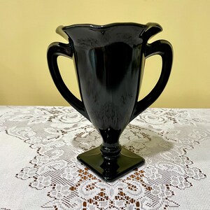 Peut inclure: Un vase en verre noir brillant dot&eacute; d'un large bord festonn&eacute;, de deux anses lat&eacute;rales incurv&eacute;es et d'un corps effil&eacute; qui descend vers une base carr&eacute;e sur pi&eacute;douche. La surface du verre est lisse et r&eacute;fl&eacute;chissante, captant la lumi&egrave;re.