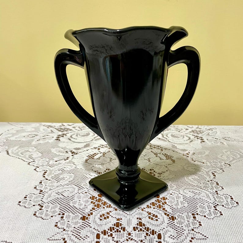 Peut inclure: Un vase en verre noir brillant dot&eacute; d'un large bord festonn&eacute;, de deux anses lat&eacute;rales incurv&eacute;es et d'un corps effil&eacute; qui descend vers une base carr&eacute;e sur pi&eacute;douche. La surface du verre est lisse et r&eacute;fl&eacute;chissante, captant la lumi&egrave;re.