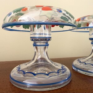 May include: Two clear glass candle holders with a mushroom-shaped top. The tops are decorated with colorful floral designs and outlined with a blue rim. The bases are clear glass with blue accents.