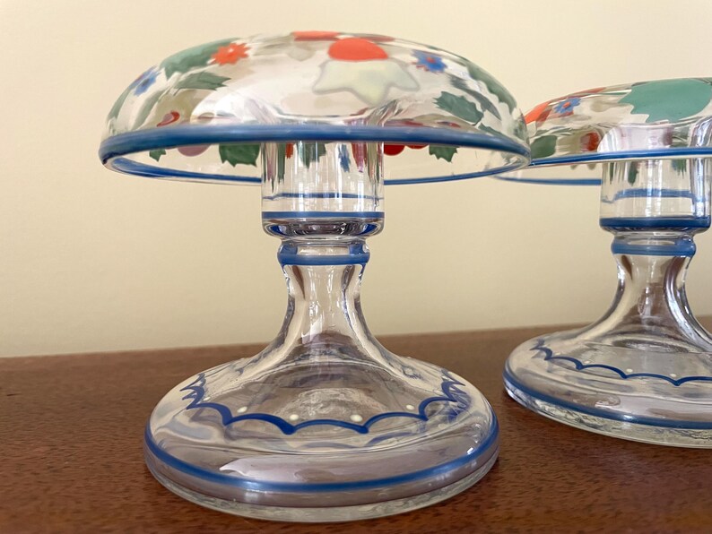May include: Two clear glass candle holders with a mushroom-shaped top. The tops are decorated with colorful floral designs and outlined with a blue rim. The bases are clear glass with blue accents.