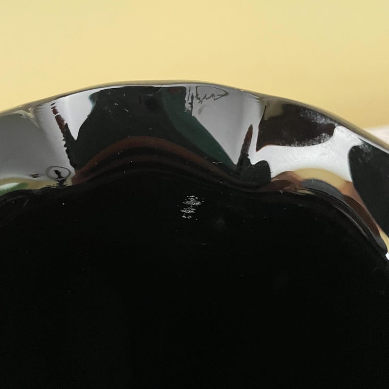 Peut inclure: Un bol en verre noir brillant pr&eacute;sentant un bord festonn&eacute; ou ondul&eacute; caract&eacute;ristique. La surface du bol est lisse et r&eacute;fl&eacute;chissante, avec des reflets lumineux le long du bord incurv&eacute;. La couleur noire profonde est uniforme sur toute la pi&egrave;ce.