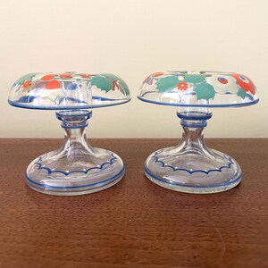 May include: Two clear glass candle holders with a floral design on the top. The floral design includes red, white, and green flowers. The candle holders have a blue trim and are on a brown surface.