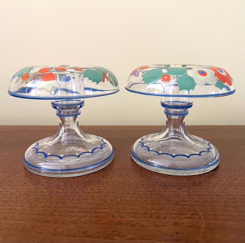 May include: Two clear glass candle holders with a floral design on the top. The floral design includes red, white, and green flowers. The candle holders have a blue trim and are on a brown surface.