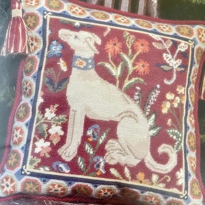 May include: A square needlepoint pillow featuring a cream-colored dog wearing a blue collar, centered on a deep red background with floral motifs. The pillow is bordered by a geometric pattern and finished with red twisted cord trim and a tassel.