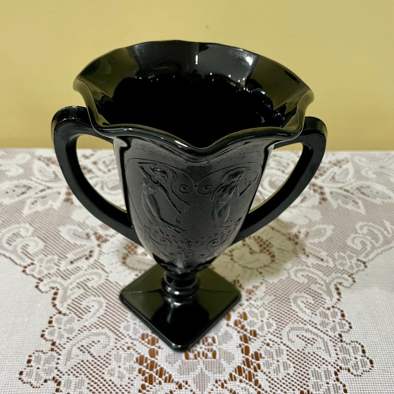 Peut inclure: Un vase en verre noir dot&eacute; d'un bord large et ondul&eacute; ainsi que de deux anses lat&eacute;rales. Le corps du vase est orn&eacute; de figures en relief et de motifs de volutes. Il repose sur une base carr&eacute;e, pr&eacute;sentant une finition sombre et brillante.