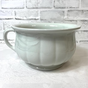 May include: A white ceramic chamber pot featuring a rounded body with vertical fluted detailing, a wide rim, and a single side handle. The vessel has a smooth, glossy finish and sits on a circular base, presenting a classic, functional design.