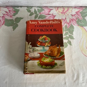 May include: A vintage cookbook titled "Amy Vanderbilt's Complete Cookbook" with a red and brown cover. The cover features illustrations of food, including a roasted turkey, fruit, and a molded salad. The book is on a floral tablecloth.