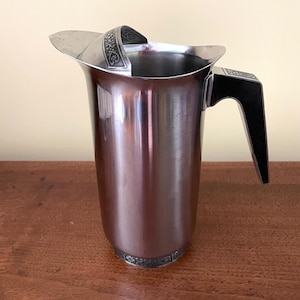 May include: A stainless steel pitcher with a polished finish. The pitcher has a black handle and decorative silver trim around the base and handle. The pitcher is designed with a flared spout and a decorative handle.