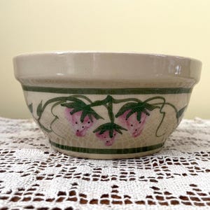 May include: A cream-colored ceramic bowl with a green and white floral design featuring pink strawberries. The bowl has a slightly rounded rim and a smooth, glazed finish.