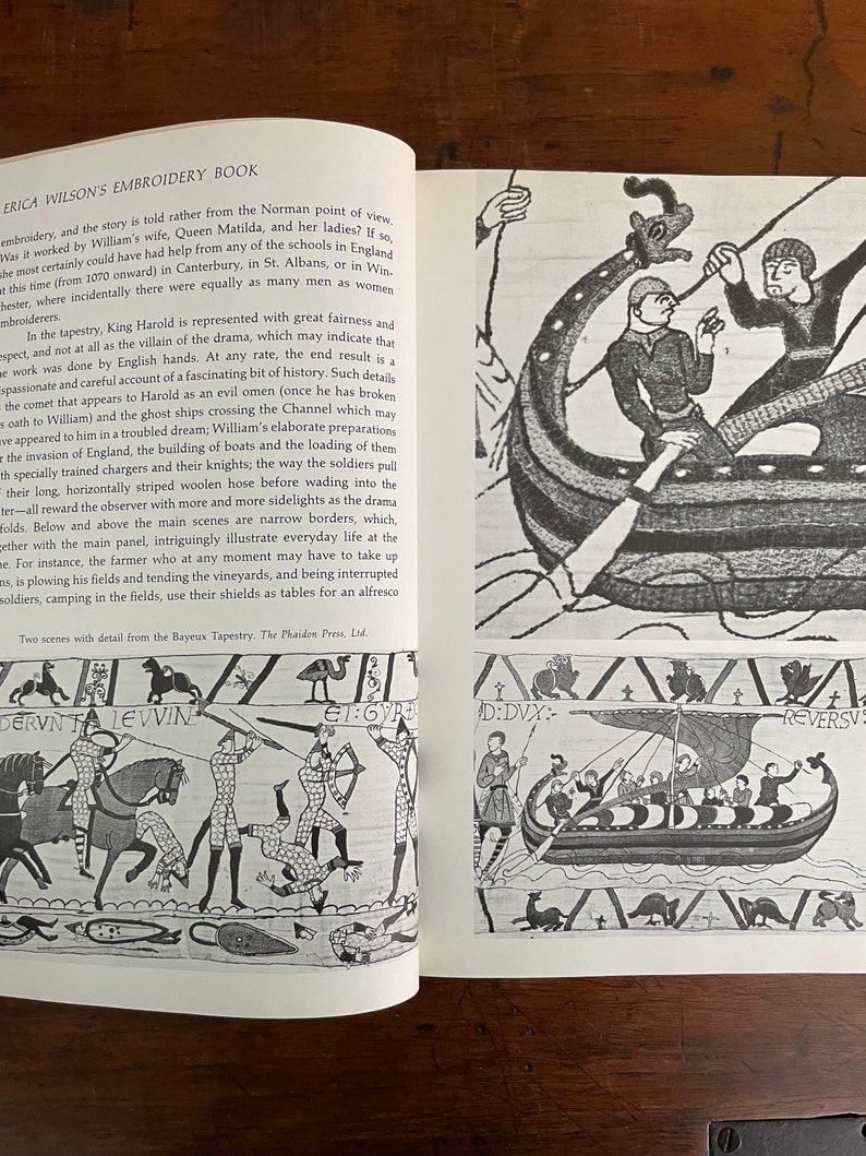 May include: Open book with a detailed illustration of the Bayeux Tapestry. The black and white drawings depict scenes of historical battles, boats, and figures. Text on the page includes the title "ERICA WILSON'S EMBROIDERY BOOK" and descriptive text about the tapestry.
