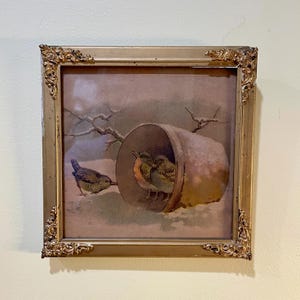May include: A square wall art piece featuring a print of three small birds in a snowy landscape. Two birds are sheltered inside a terracotta-colored pot, while one stands outside. The artwork is encased in a metallic gold frame with ornate corner details.