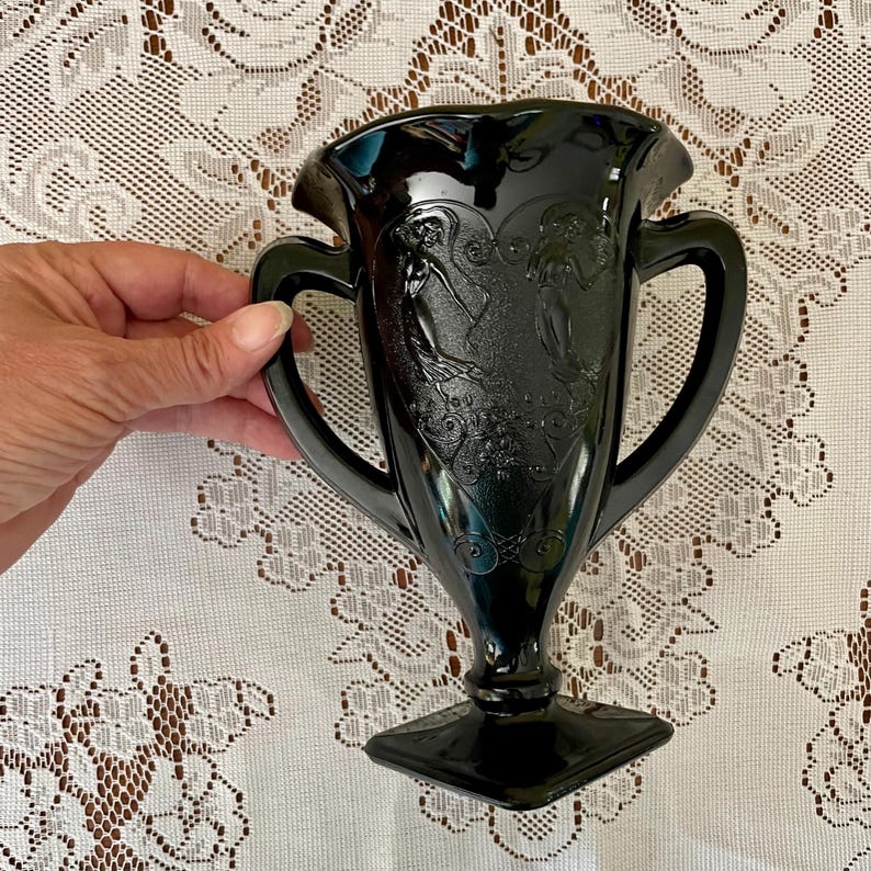 May include: A black glass vase featuring two handles and a square base. The body of the vase displays a raised relief design of two figures in flowing garments surrounded by decorative scrollwork patterns on a textured, heart-shaped background.