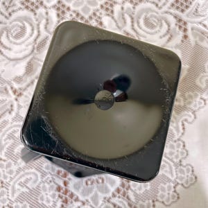 Peut inclure: Un objet carr&eacute;, noir et brillant, aux coins arrondis et dot&eacute; d'une indentation circulaire au centre. La surface pr&eacute;sente de fines rayures visibles et une finition r&eacute;fl&eacute;chissante, avec une petite ouverture circulaire sombre situ&eacute;e au milieu.