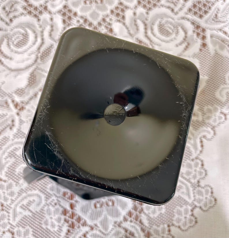Peut inclure: Un objet carr&eacute;, noir et brillant, aux coins arrondis et dot&eacute; d'une indentation circulaire au centre. La surface pr&eacute;sente de fines rayures visibles et une finition r&eacute;fl&eacute;chissante, avec une petite ouverture circulaire sombre situ&eacute;e au milieu.