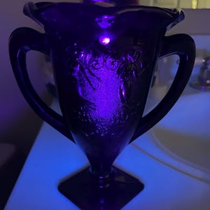 Peut inclure: Un vase en verre violet fonc&eacute; avec deux anses et une base carr&eacute;e. La surface pr&eacute;sente un relief repr&eacute;sentant deux personnages et des motifs floraux, &eacute;clair&eacute; par une lumi&egrave;re violette vive qui souligne les textures et les d&eacute;tails complexes.