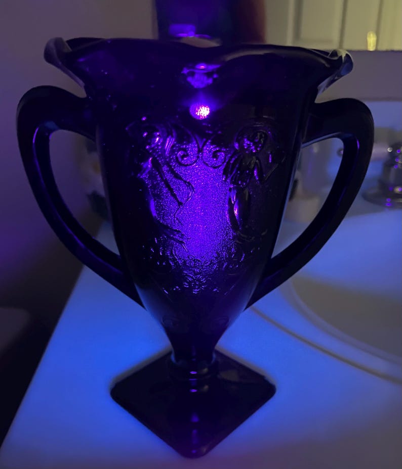 Peut inclure: Un vase en verre violet fonc&eacute; avec deux anses et une base carr&eacute;e. La surface pr&eacute;sente un relief repr&eacute;sentant deux personnages et des motifs floraux, &eacute;clair&eacute; par une lumi&egrave;re violette vive qui souligne les textures et les d&eacute;tails complexes.
