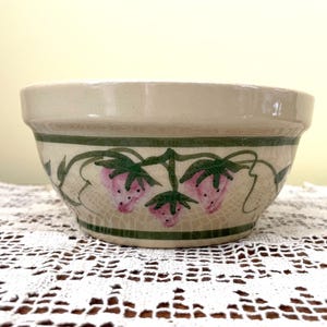 May include: A cream-colored ceramic bowl with a green and pink strawberry design. The bowl has a wide rim and a rounded bottom.