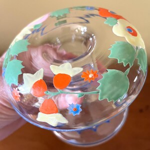 May include: A clear glass candle holder with a floral design. The flowers are painted in orange, white, and blue, with green leaves. The candle holder has a round base and a flared top.