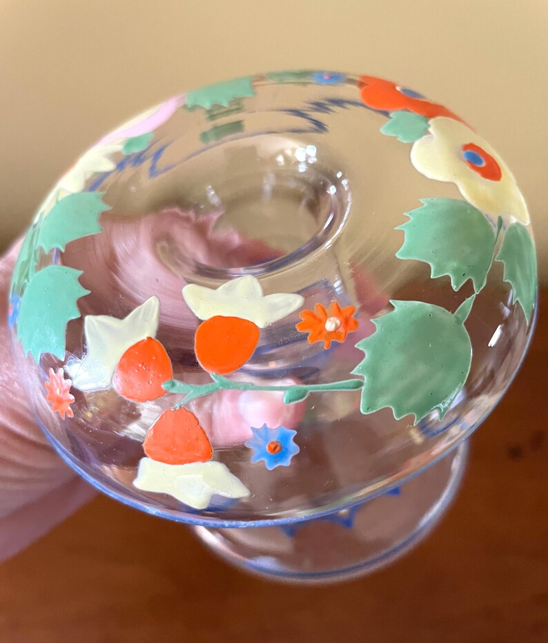 May include: A clear glass candle holder with a floral design. The flowers are painted in orange, white, and blue, with green leaves. The candle holder has a round base and a flared top.