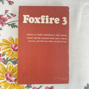 May include: A vintage book cover with the title "Foxfire 3" in white letters on a reddish-orange background. The cover lists topics such as animal care, wild plant foods, and plain living. The book is on a floral patterned surface.