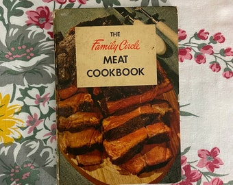 May include: Vintage cookbook titled "The Family Circle Meat Cookbook" with a beige cover and a picture of cooked meat. The book is set against a floral patterned tablecloth with pink, gray, and yellow flowers.
