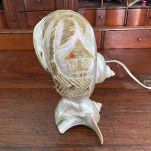 May include: A pearlescent seashell lamp with a carved design of a house. The lamp is made from a large conch shell, with a smaller shell forming the base. The lamp has a white cord and is designed to sit on a table or desk.