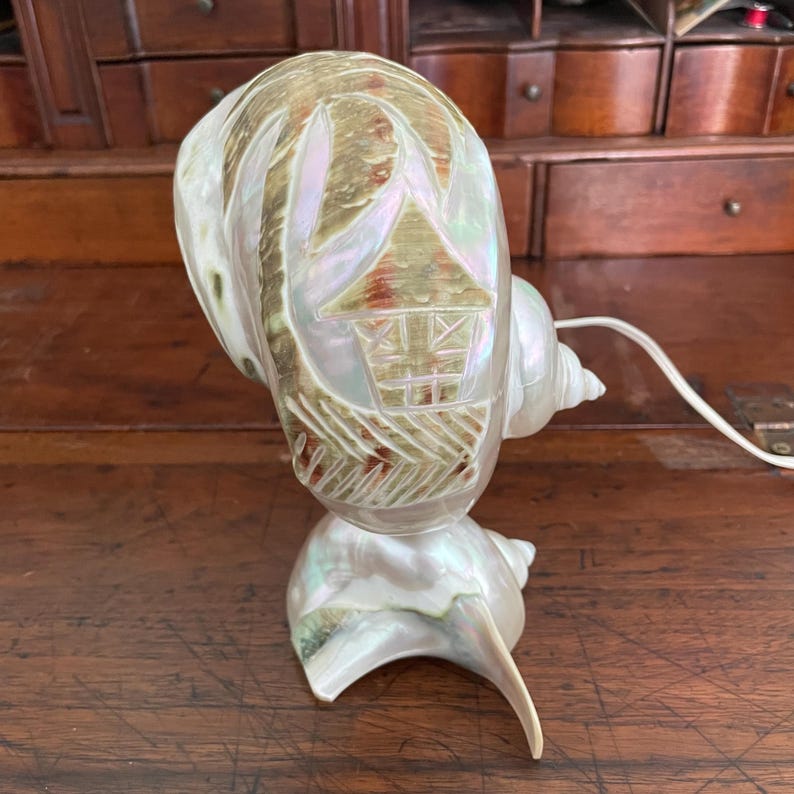 May include: A pearlescent seashell lamp with a carved design of a house. The lamp is made from a large conch shell, with a smaller shell forming the base. The lamp has a white cord and is designed to sit on a table or desk.