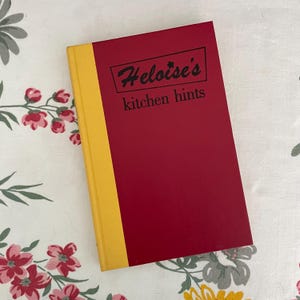 May include: A hardcover book titled Heloise's kitchen hints with a deep red front cover and a bright yellow spine. The title is printed in black text inside a rectangular border, with the words kitchen hints appearing directly below the box.