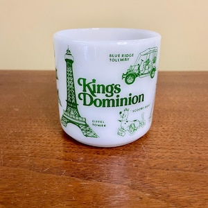 May include: A white milk glass mug featuring green printed graphics of the Eiffel Tower, a vintage car labeled Blue Ridge Tollway, and the cartoon character Scooby Doo. The text Kings Dominion is printed in the center in a bold green font.