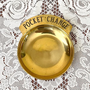 May include: A vintage brass pocket change dish with the words "POCKET CHANGE" in black lettering. The dish is round with a curved top and a shiny, golden finish. The dish is likely used to hold coins or small items.