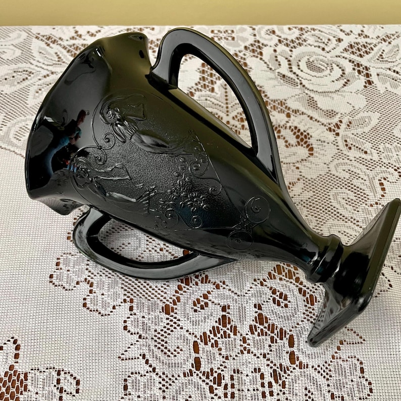 Peut inclure: Un pichet en verre noir brillant orn&eacute; d'un relief repr&eacute;sentant une silhouette aux cheveux flottants et des motifs floraux tourbillonnants. Le r&eacute;cipient pr&eacute;sente un bord &eacute;vas&eacute;, une base sur pied robuste et deux anses incurv&eacute;es.